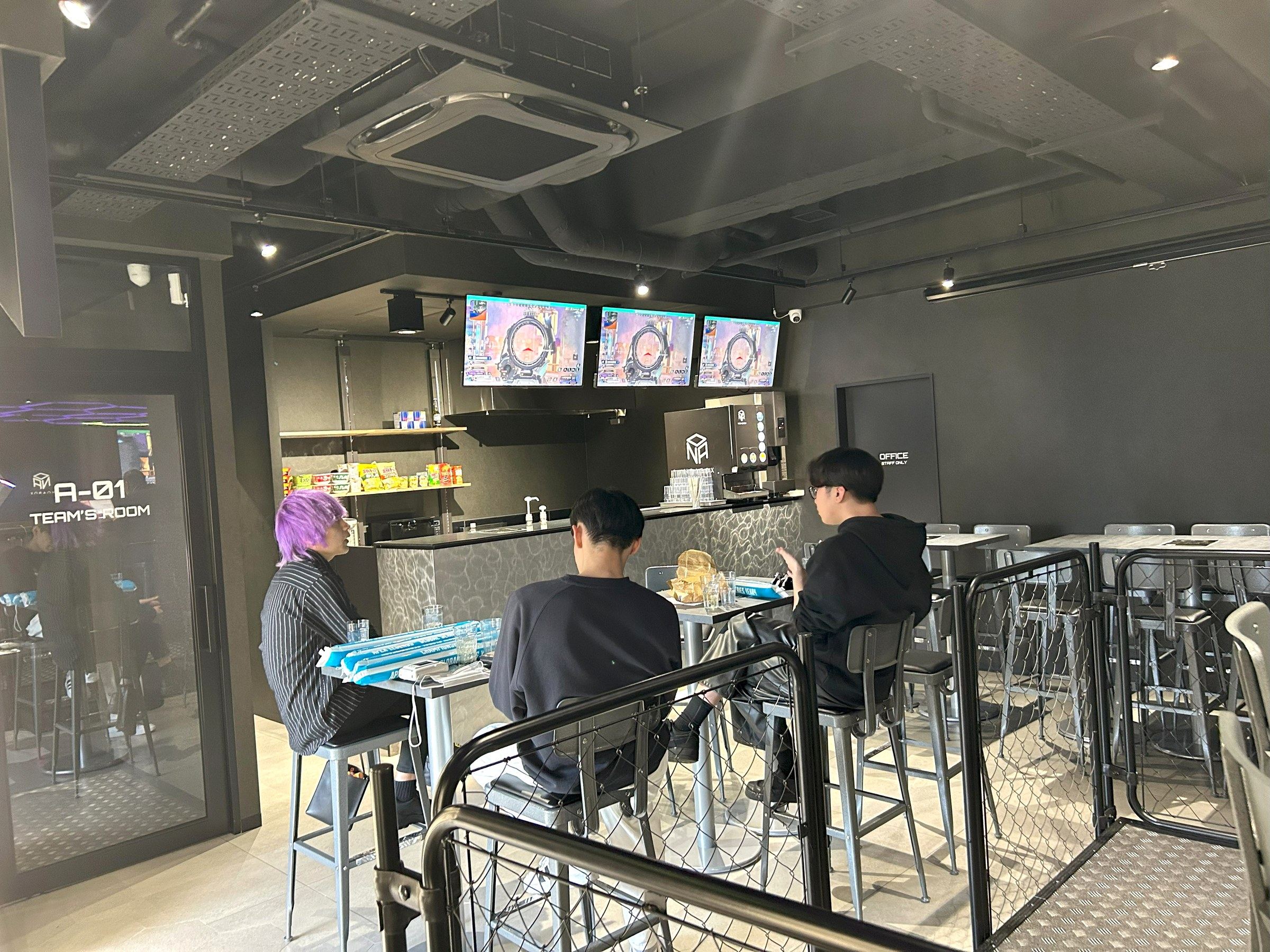 カフェスペースのハイテーブルに座る3人。奥にカウンターと、飲み物やお菓子が並ぶ棚が見える